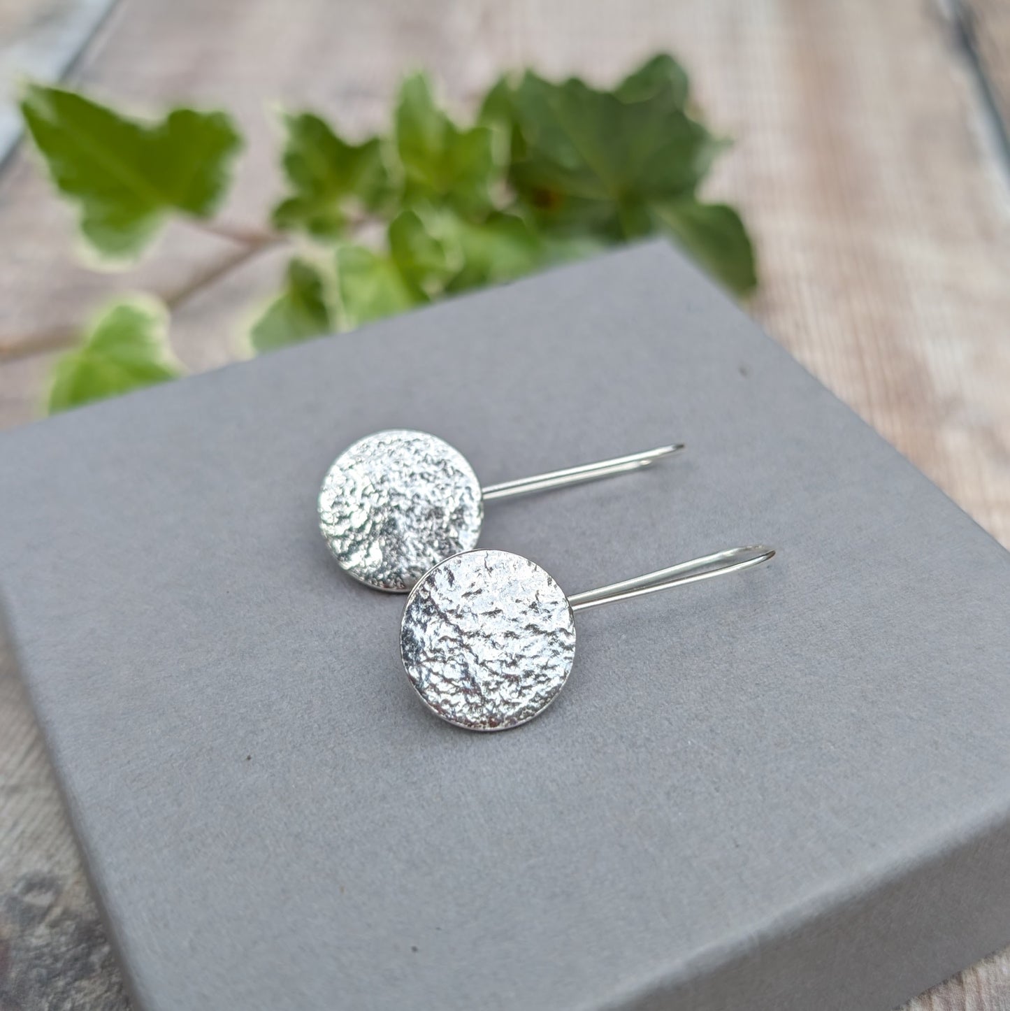 Silver organic disc long earrings handmade from recycled Sterling Silver, with textured circular discs resting on a grey jewellery box.