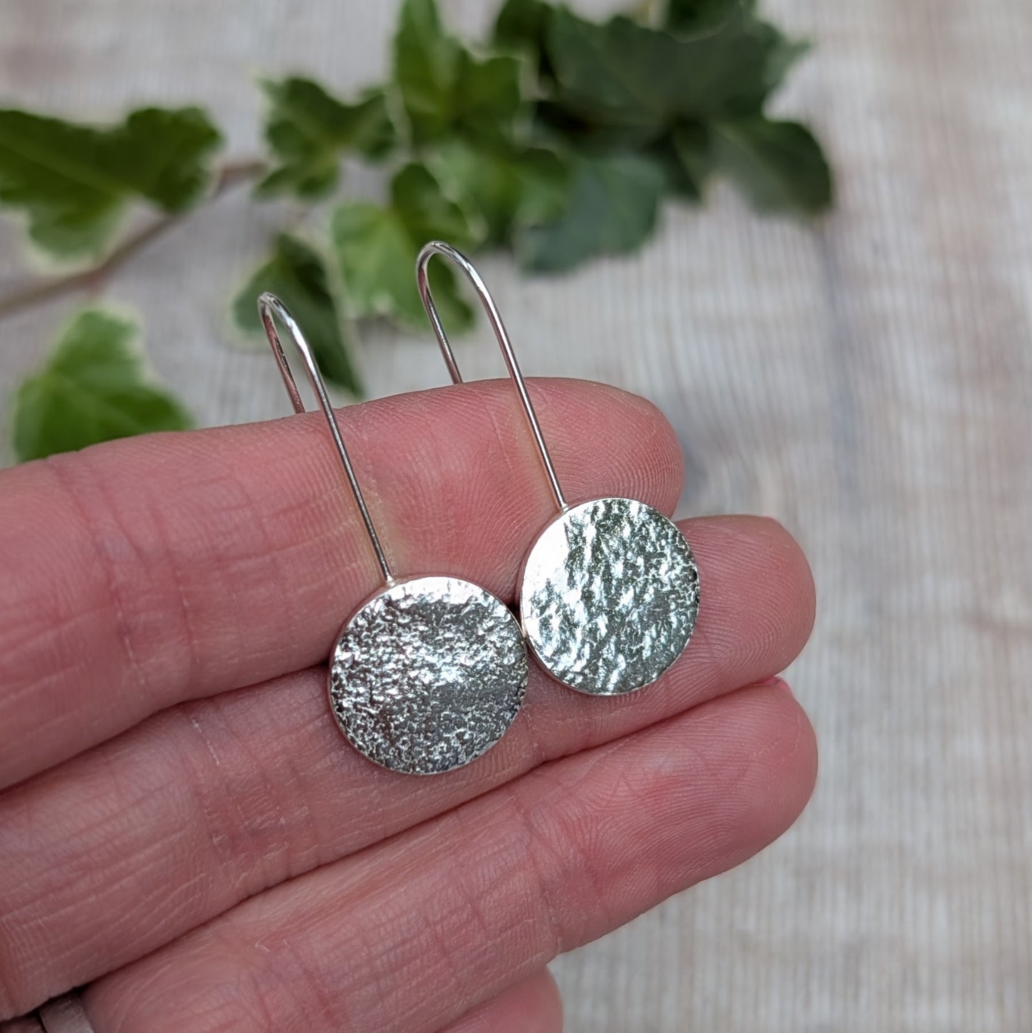 Silver organic disc long earrings held in a hand, showing the textured round silver discs and long curved ear wires.