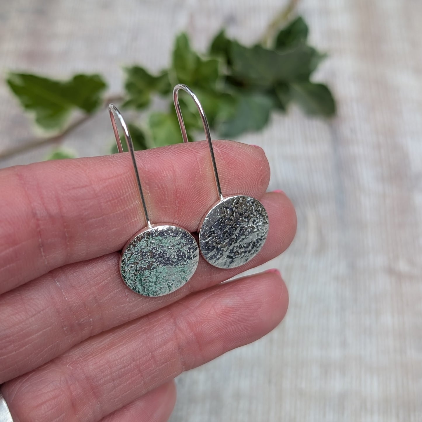 Close up of silver organic disc long earrings held in a hand, showing the textured surface of each round silver disc.
