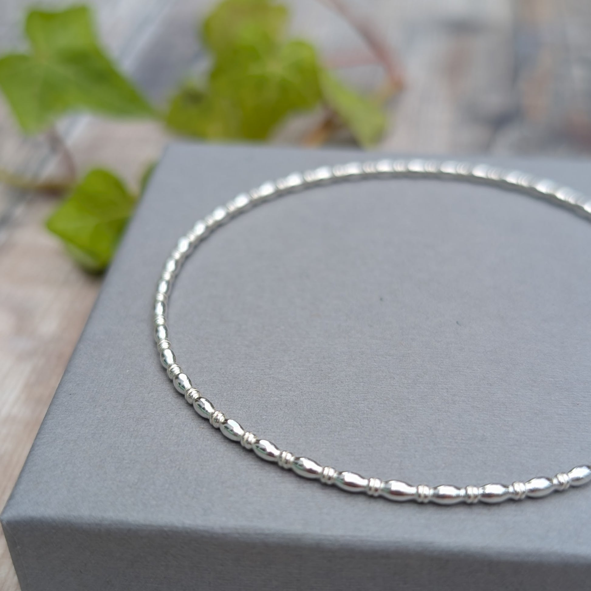 Close up of the Silver Oval Beaded Wire Round Bangle handmade from recycled Sterling Silver 2mm square wire, resting on a grey jewellery box with the oval beaded pattern in focus.