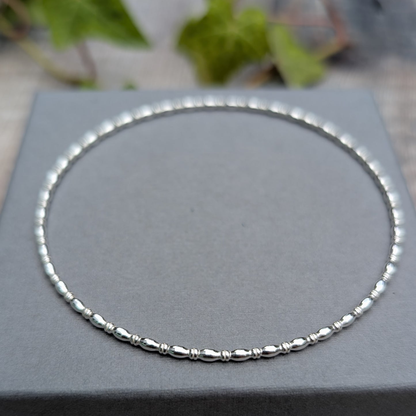 Silver Oval Beaded Wire Round Bangle handmade from recycled Sterling Silver 2mm square wire, shown flat on a grey jewellery box.