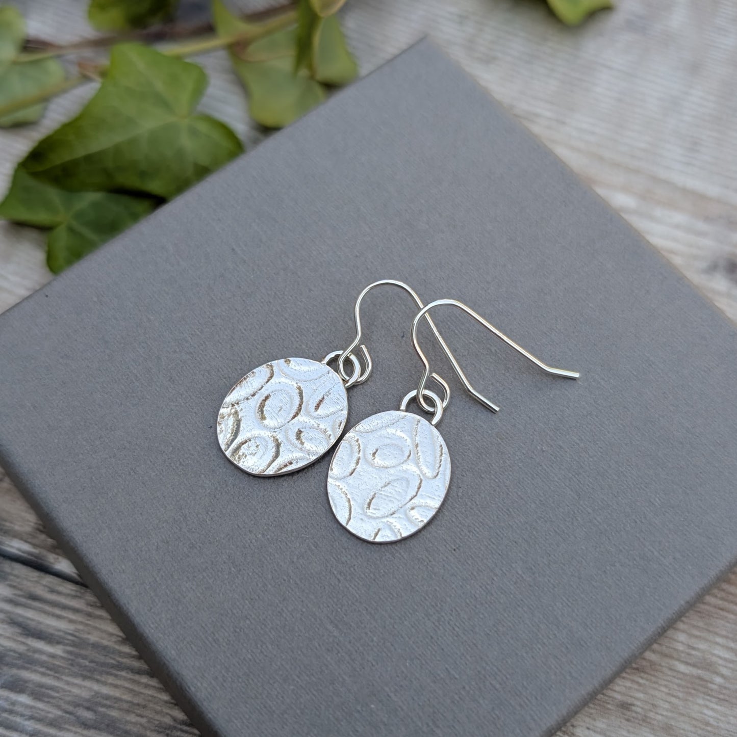 Silver Oval Disc Earrings with Oval Pattern displayed on a grey box with natural background