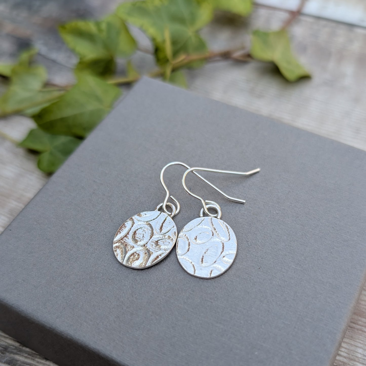 Silver Oval Disc Earrings with Oval Pattern shown close up on a grey box outdoors