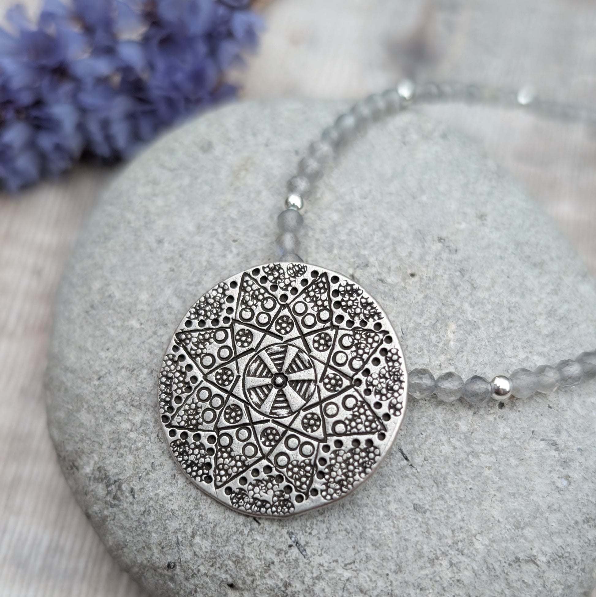 A close-up of the silver patterned disc necklace with Labradorite resting on a grey stone, with a bunch of purple flowers in the background. The Karen Hill Tribe silver disc shows an intricate geometric pattern.