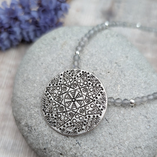 A close-up of the silver patterned disc necklace with Labradorite resting on a grey stone, with a bunch of purple flowers in the background. The Karen Hill Tribe silver disc shows an intricate geometric pattern.