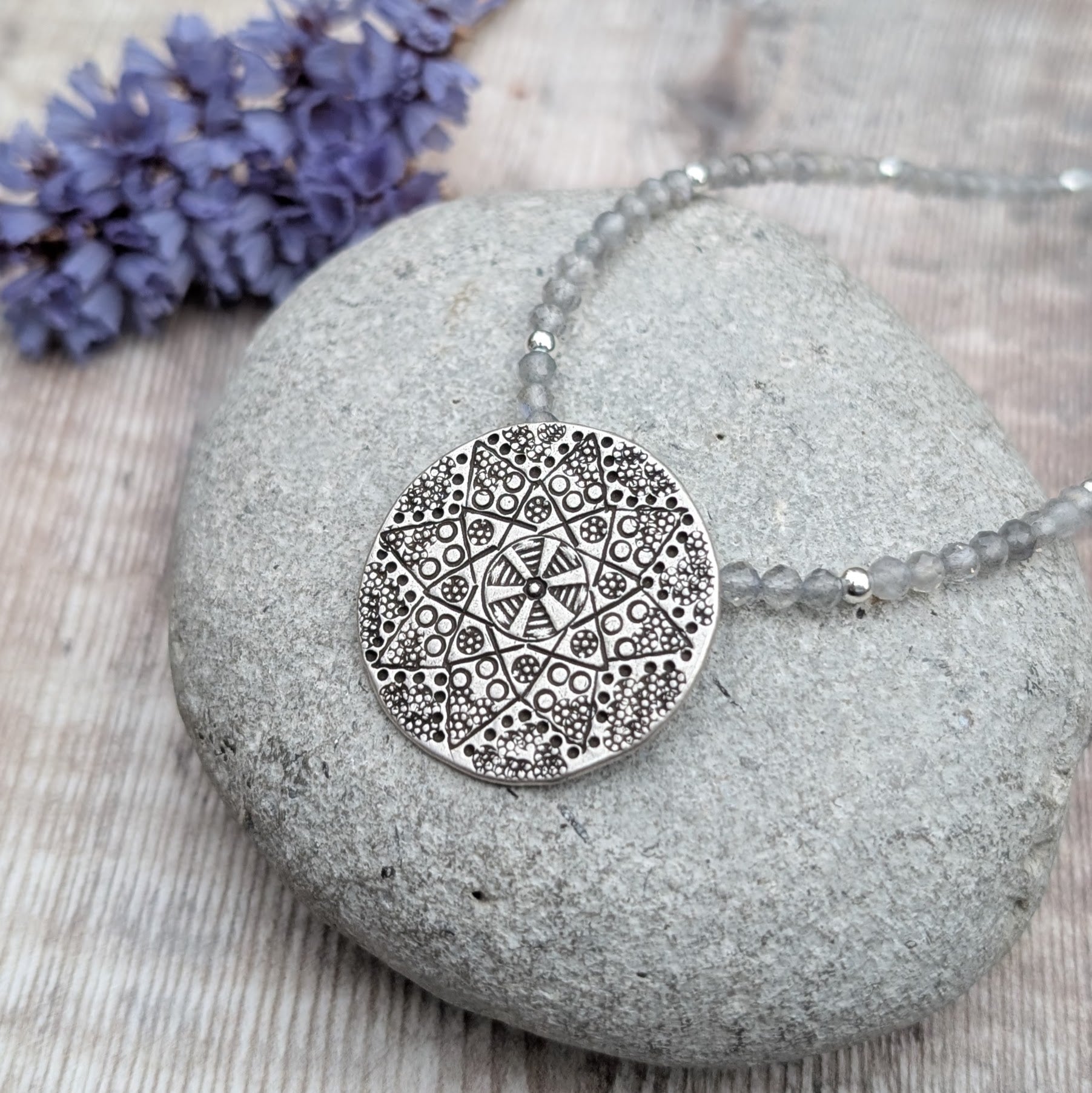 A silver patterned disc necklace with Labradorite beads laid on a smooth grey stone, with soft purple flowers blurred in the background. The disc features an intricate raised design handmade by the Karen Hill Tribe in Thailand.