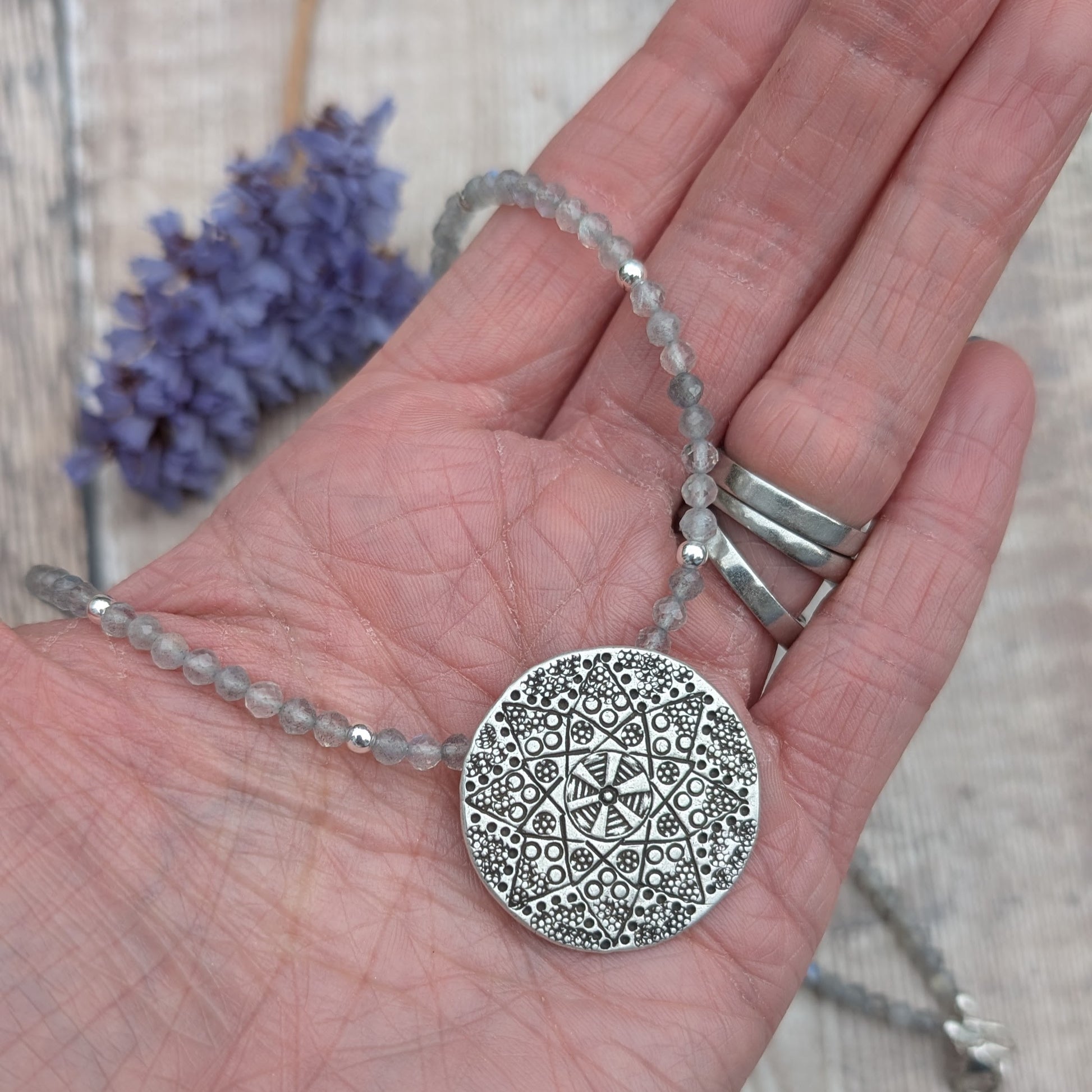 A hand holding the silver patterned disc necklace with Labradorite beads. The close-up shows the detailed raised pattern on the Karen Hill Tribe silver disc.