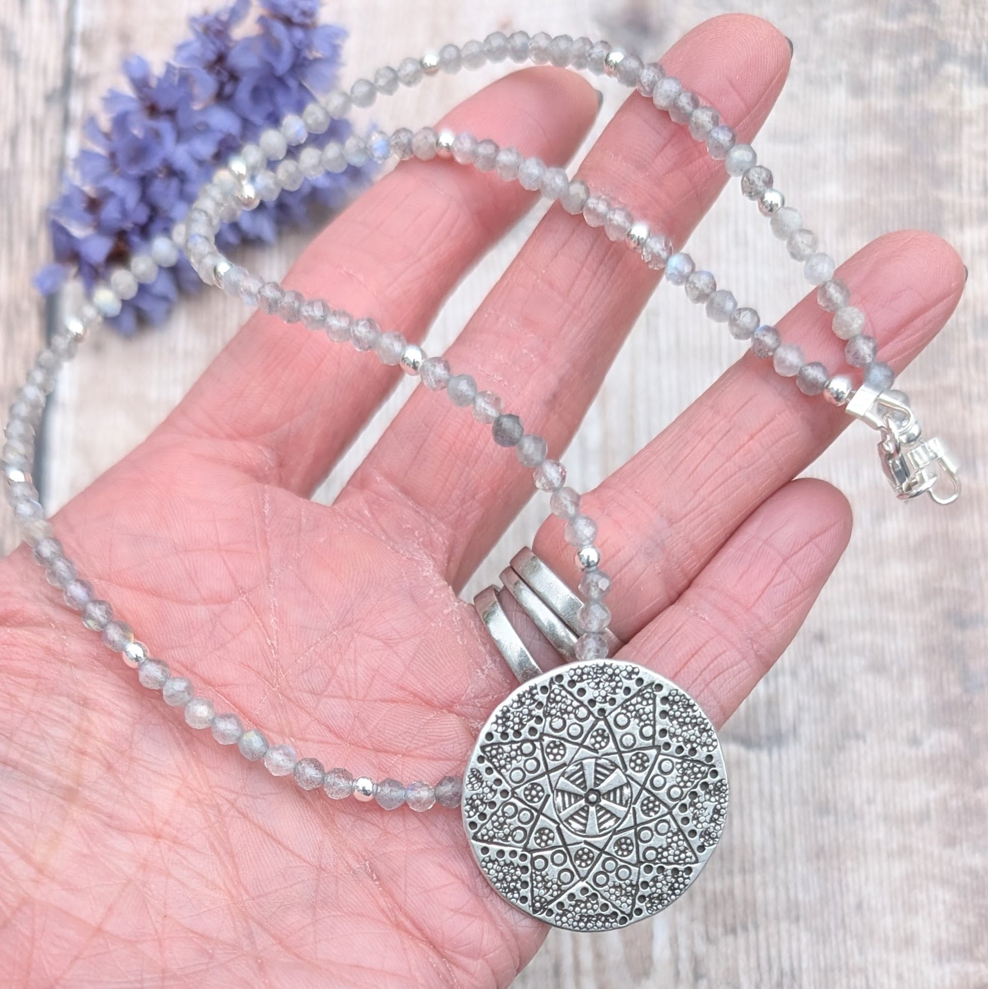 A hand holding the full length of the silver patterned disc necklace with Labradorite beads. The textured silver disc and faceted grey-blue Labradorite beads are clearly visible.