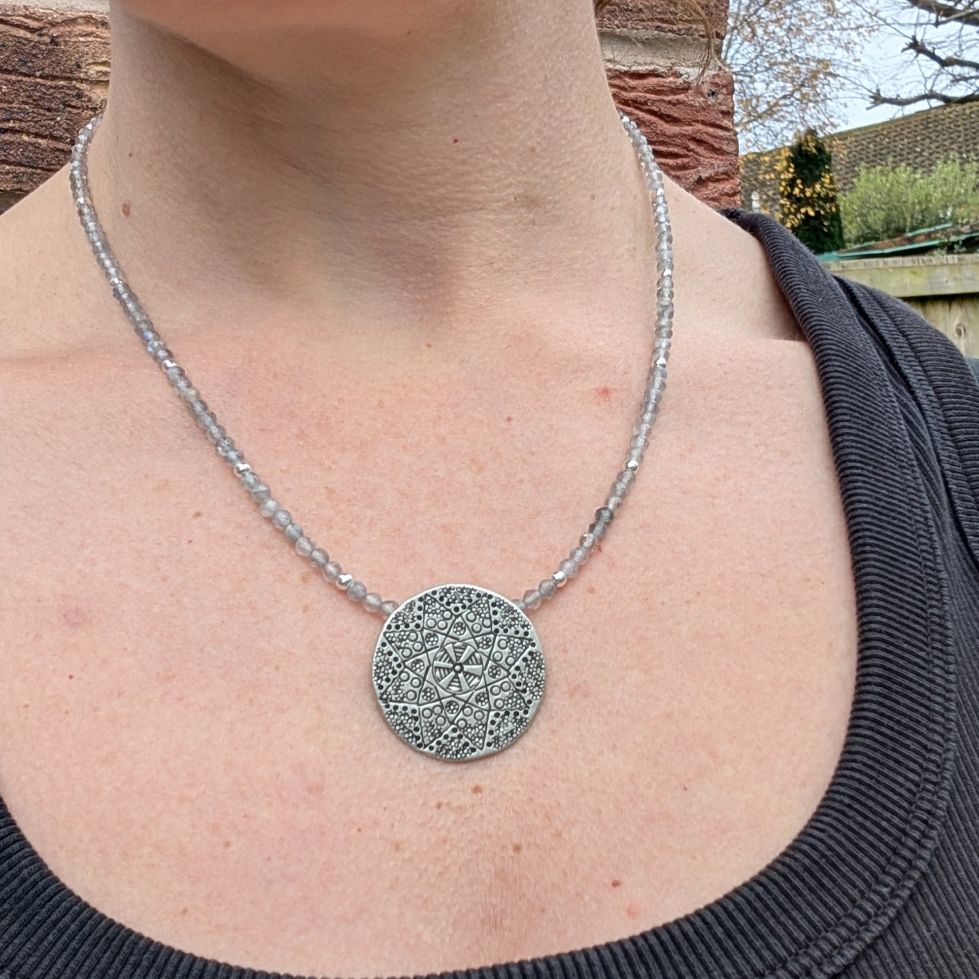 The silver patterned disc necklace with Labradorite worn around a person’s neck, showing the size and drape of the piece.