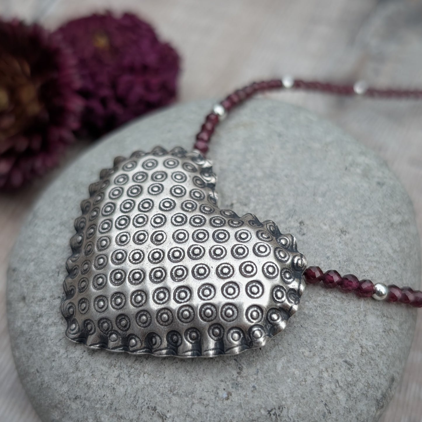 A close-up of the silver patterned heart necklace with Rhodolite Garnet resting on a grey stone, focusing on the intricate circular pattern on the heart pendant and the faceted deep-red garnet beads.