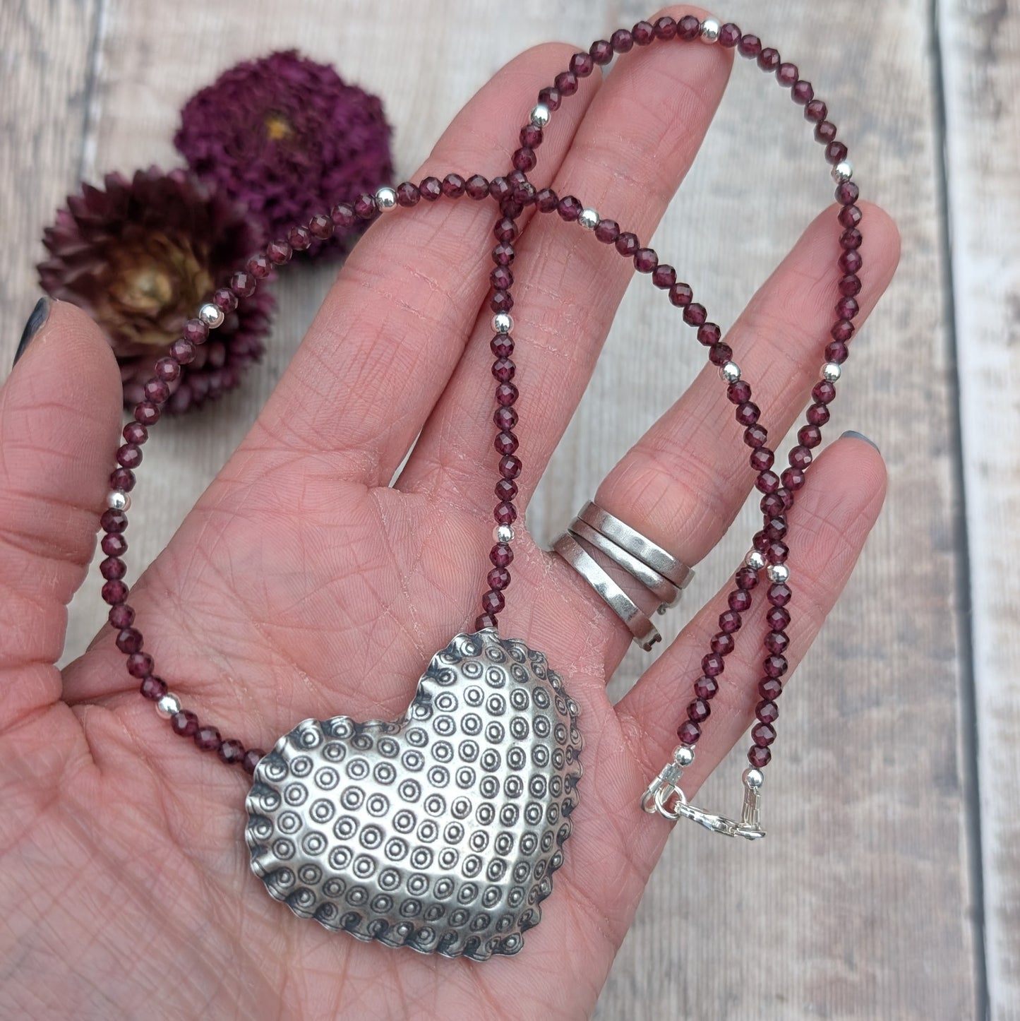 A silver patterned heart necklace with Rhodolite Garnet held in a hand. The heart pendant features a raised circular pattern, and the necklace is strung with deep berry-coloured faceted Rhodolite Garnet beads and small silver accents.