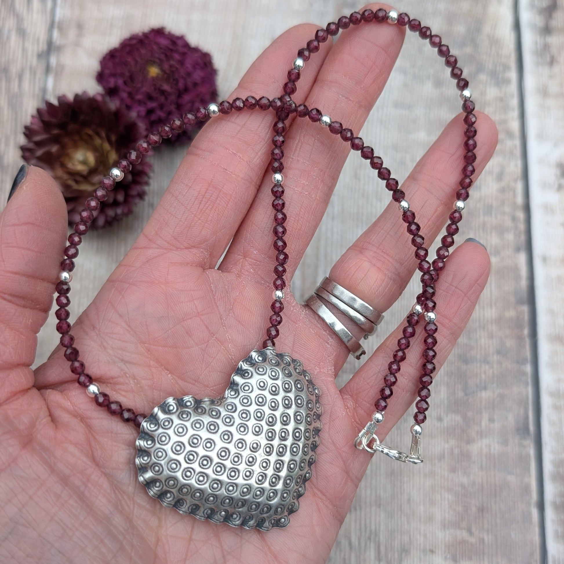 A silver patterned heart necklace with Rhodolite Garnet held in a hand. The heart pendant features a raised circular pattern, and the necklace is strung with deep berry-coloured faceted Rhodolite Garnet beads and small silver accents.