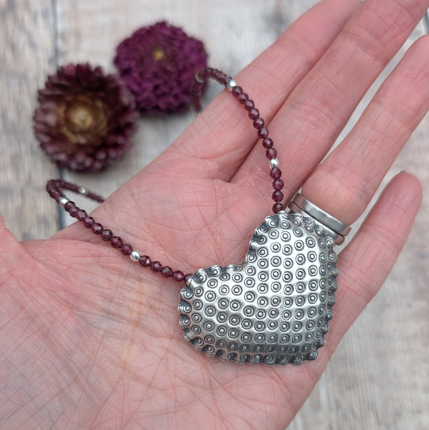A hand holding the silver patterned heart necklace with Rhodolite Garnet, showing a clear view of the textured silver heart pendant and the rich reddish-purple garnet beads.