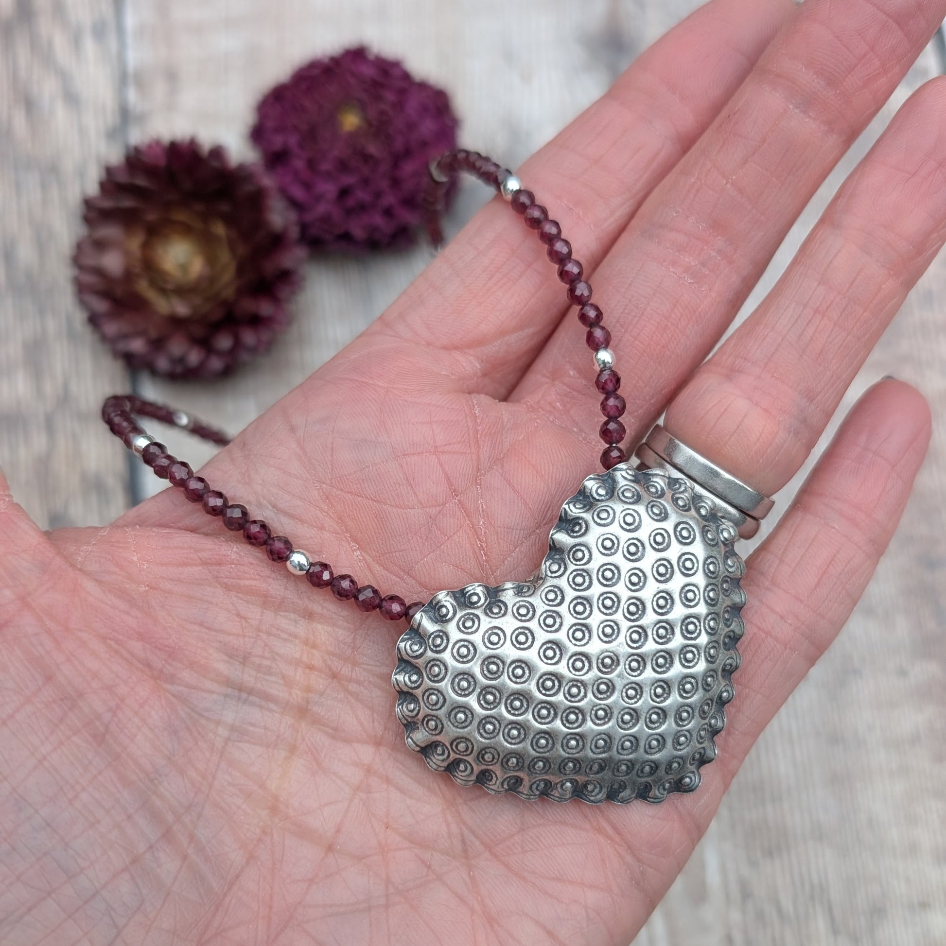 A hand holding the silver patterned heart necklace with Rhodolite Garnet, showing a clear view of the textured silver heart pendant and the rich reddish-purple garnet beads.