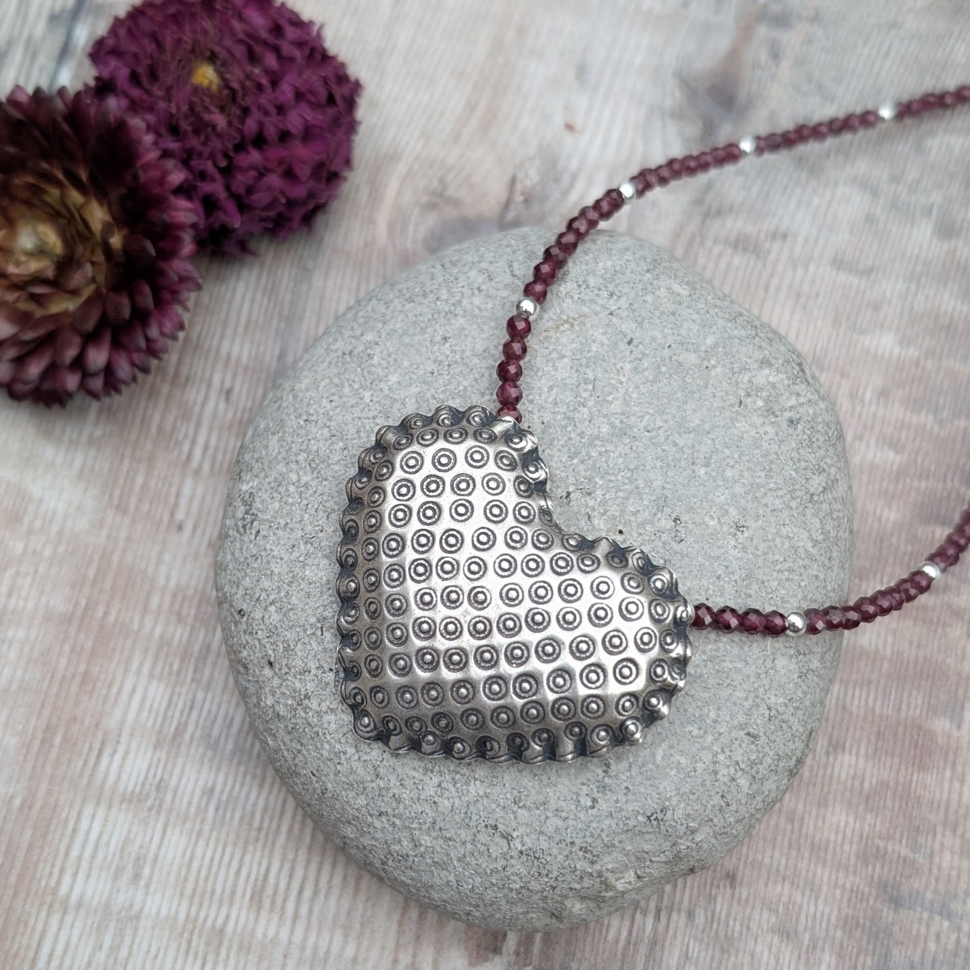 The silver patterned heart necklace with Rhodolite Garnet displayed on a smooth grey stone, with dark pink dried flowers in the background. The raised circular design on the heart pendant is visible in detail.