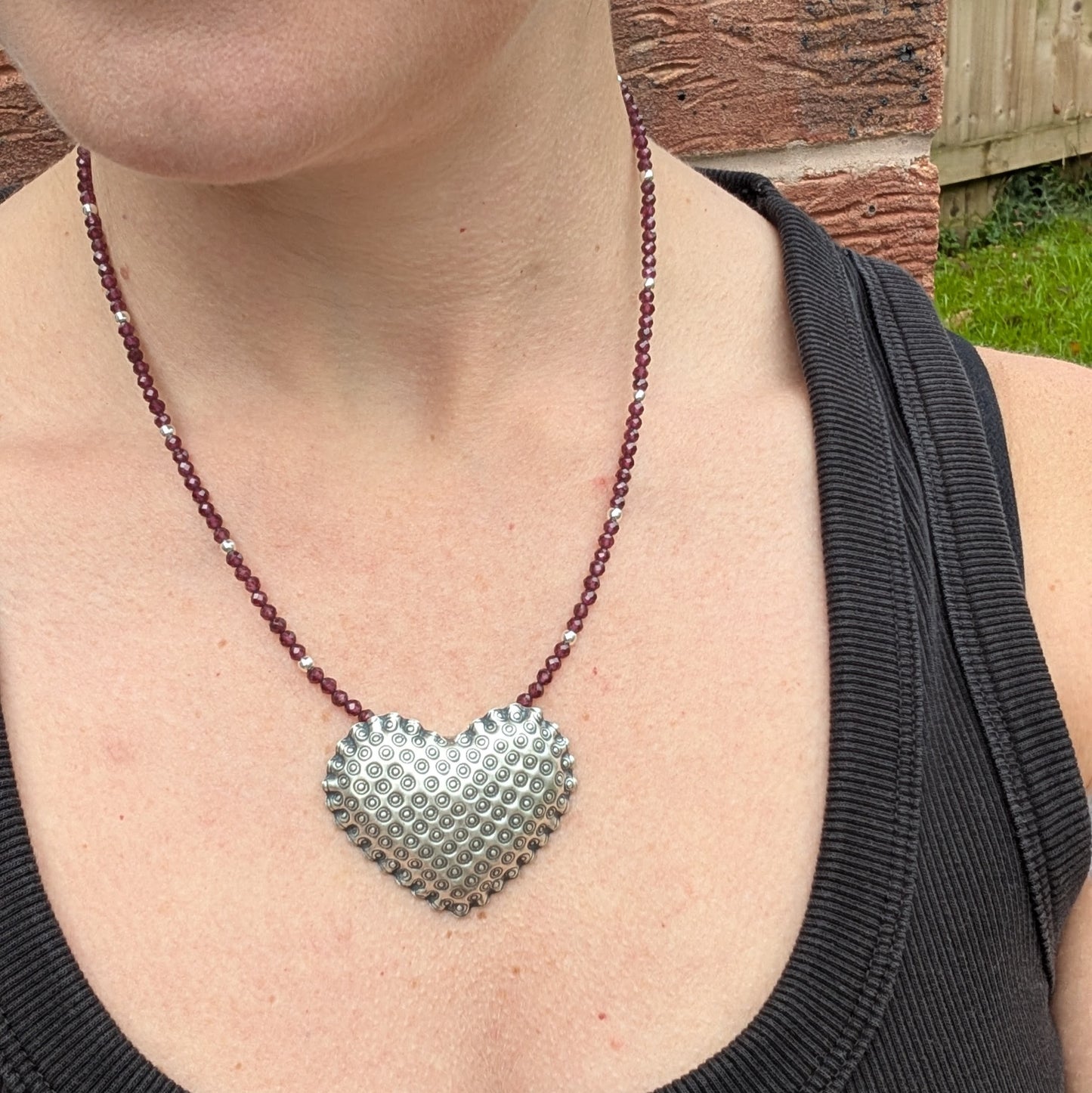 The silver patterned heart necklace with Rhodolite Garnet worn around a person’s neck, showing how the heart pendant sits when worn.