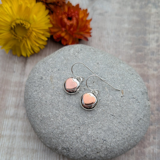 Silver pebble earrings with copper hearts resting on a smooth grey stone, with yellow and orange flowers in the background.