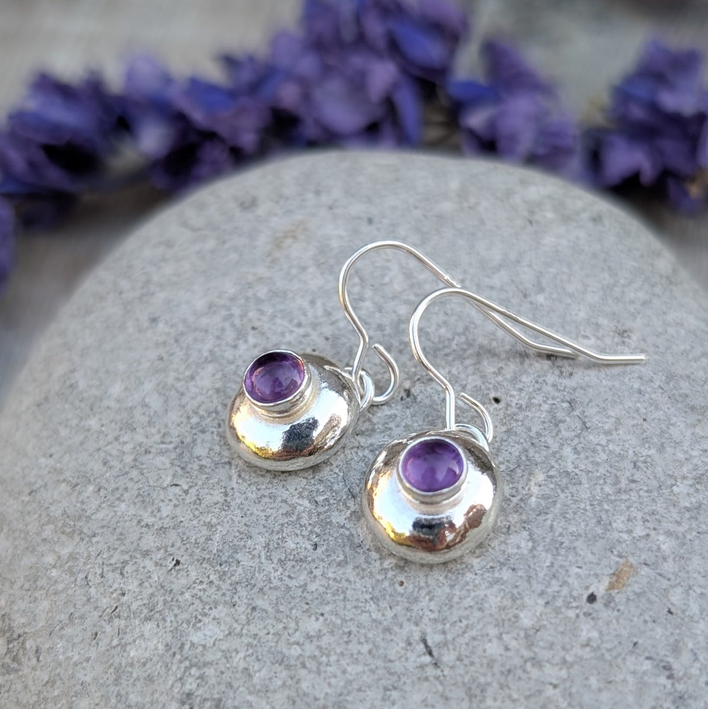 Silver pebble earrings with Amethyst resting on a stone, with purple flowers in the background.