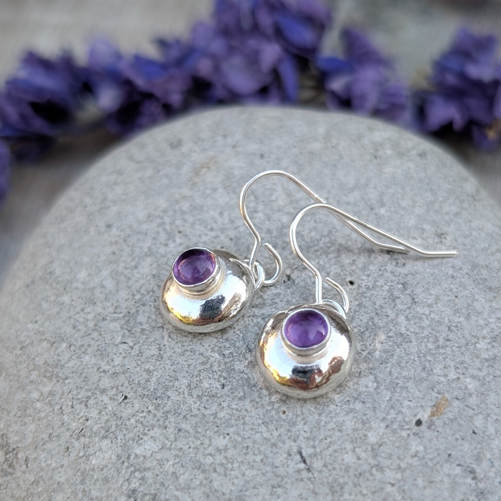 Silver pebble earrings with Amethyst resting on a stone, with purple flowers in the background.