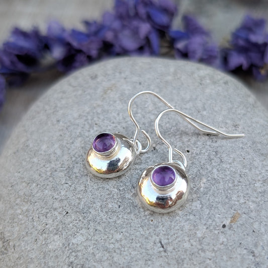 Silver pebble earrings with Amethyst resting on a stone, with purple flowers in the background.