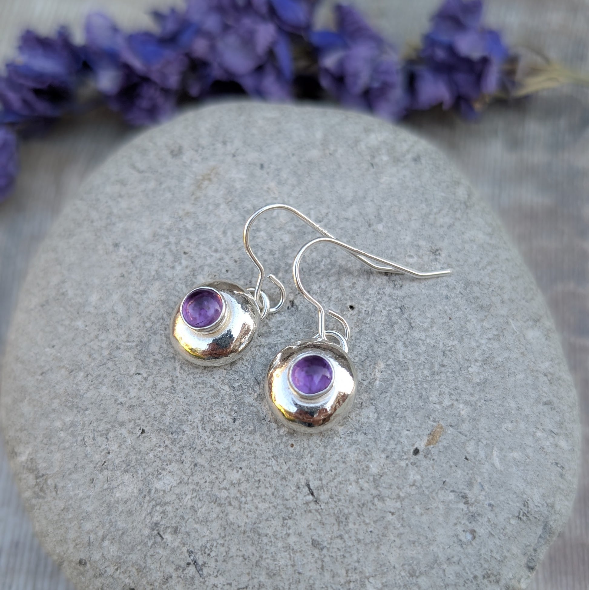 Silver pebble earrings with Amethyst placed on a smooth stone, purple flowers blurred behind.