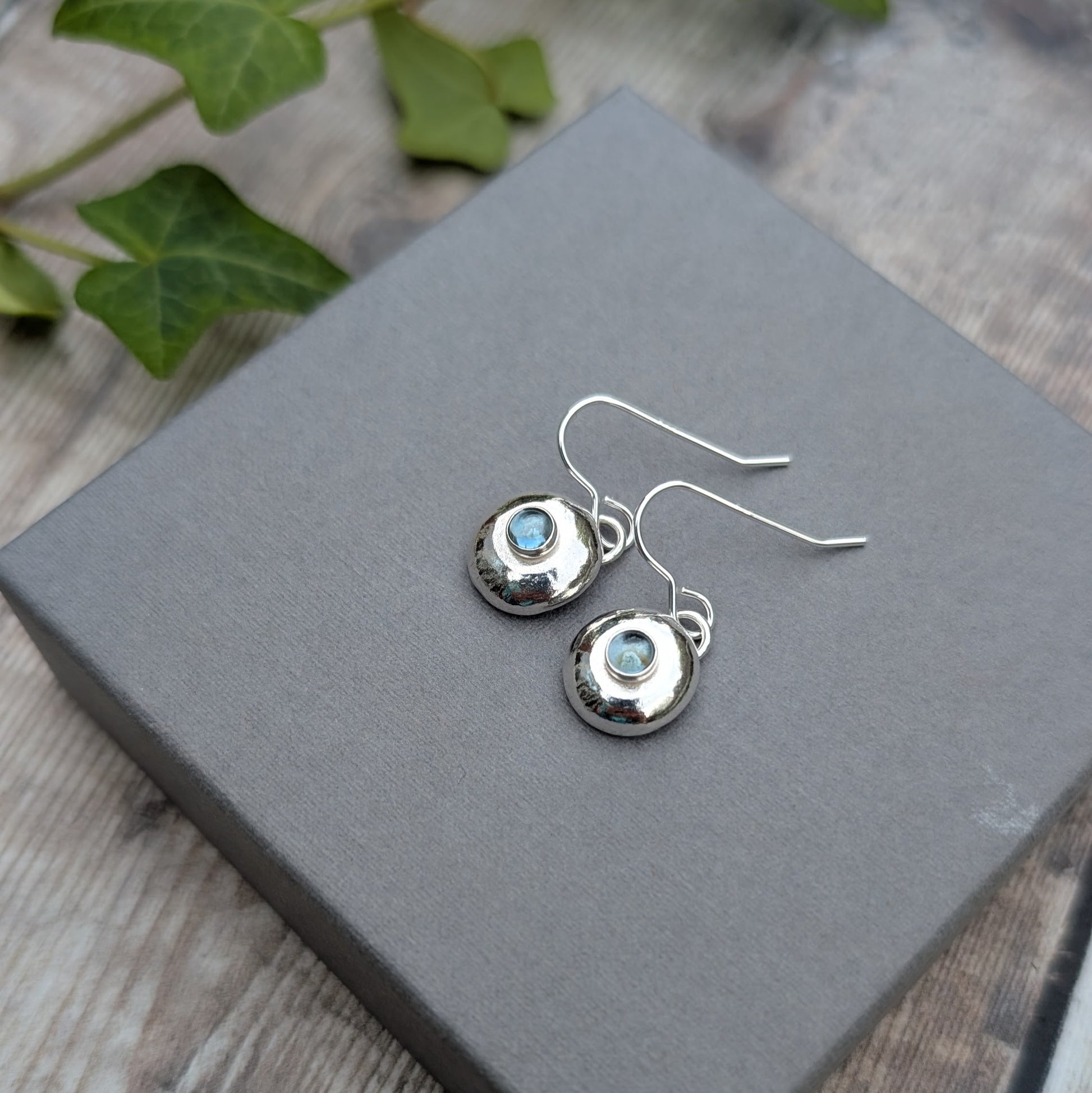 Silver Pebble Earrings with Blue Topaz on grey box with green leaves in background