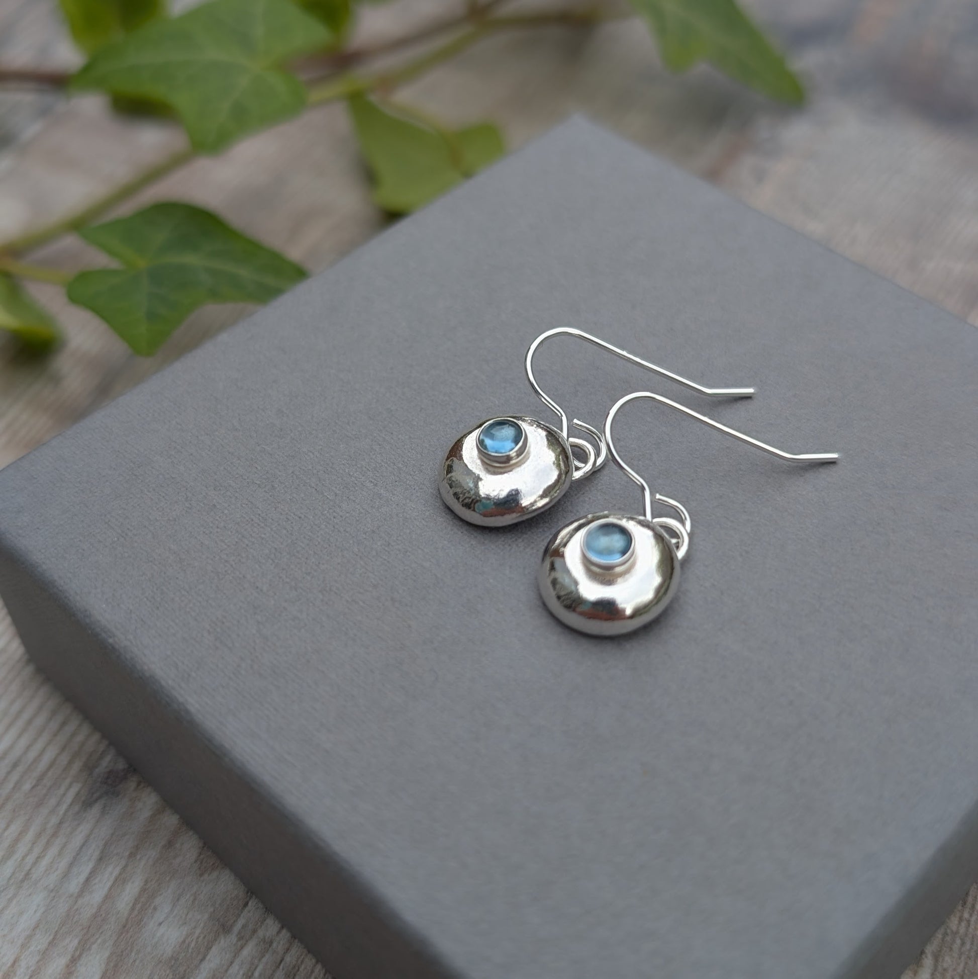 Silver Pebble Earrings with Blue Topaz resting on a grey box with soft blue gemstones in the centre