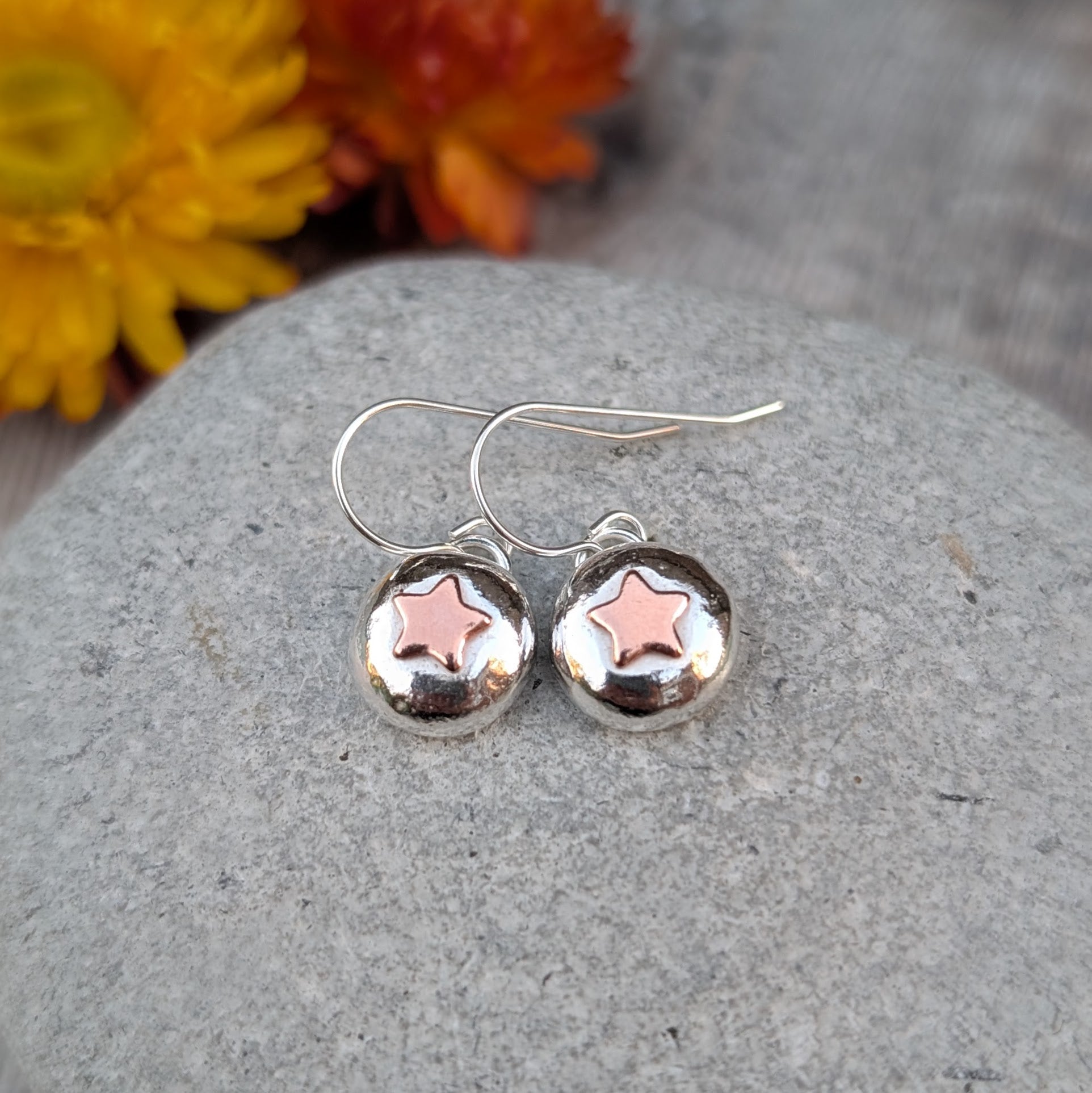 Silver pebble earrings with copper star design, photographed with warm-toned flowers in the background.