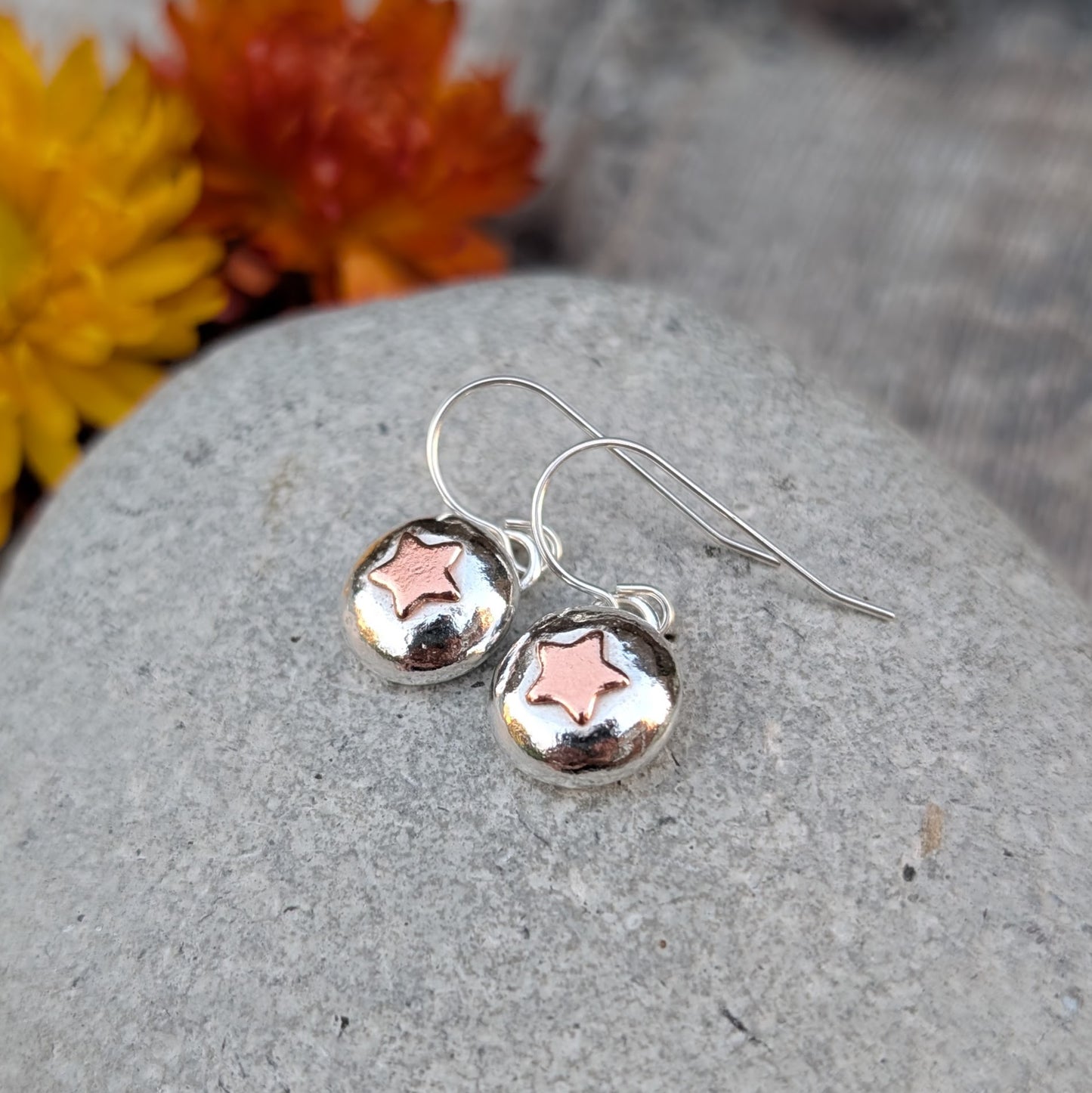 Handmade silver pebble drop earrings with small copper star details, shown resting on a grey stone.