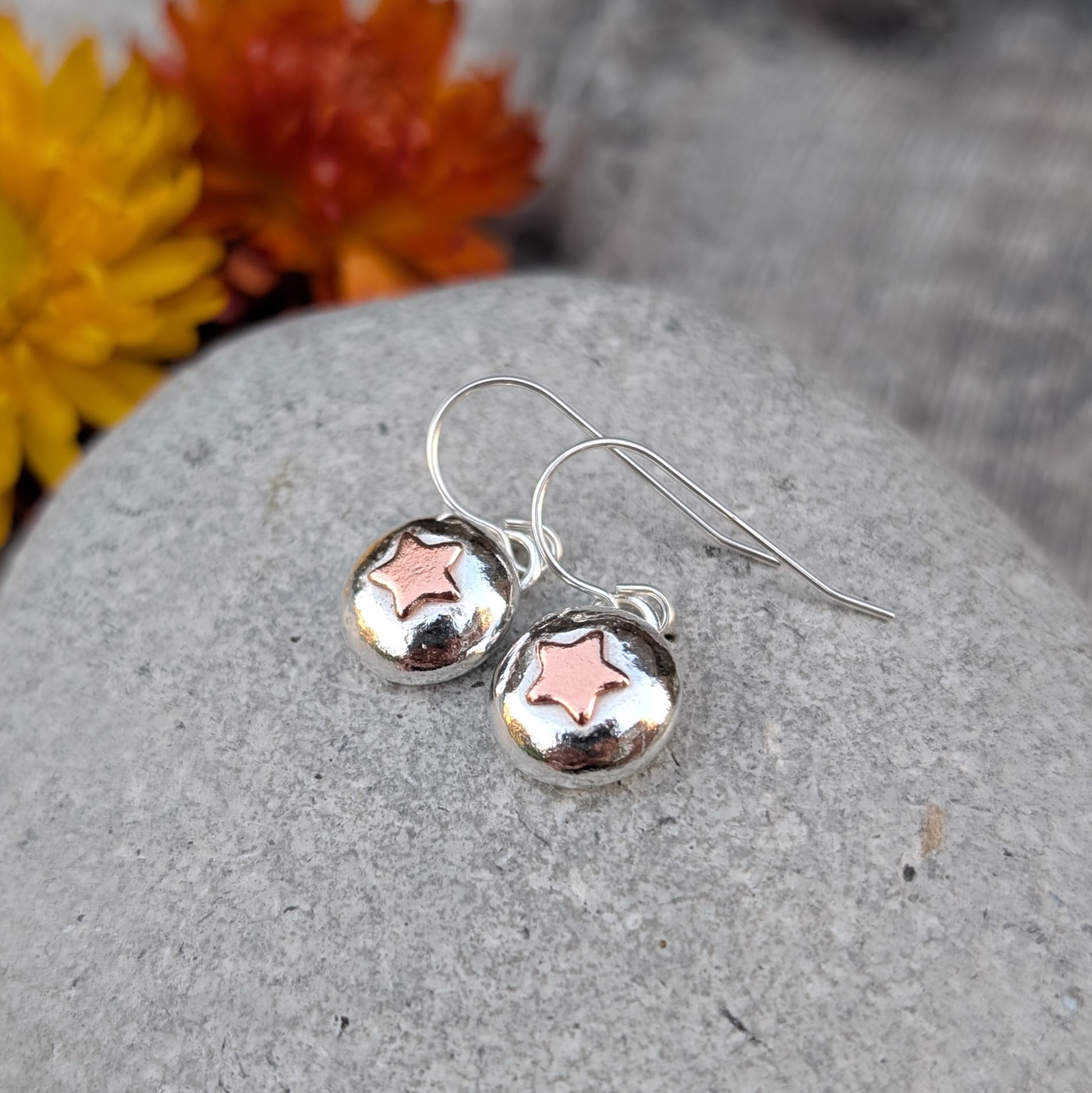 Handmade silver pebble drop earrings with small copper star details, shown resting on a grey stone.