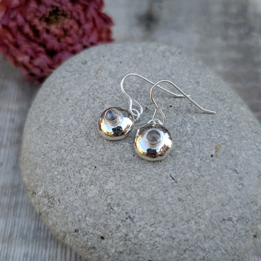 Silver pebble earrings with Labradorite resting on a stone, with a red flower in the background.