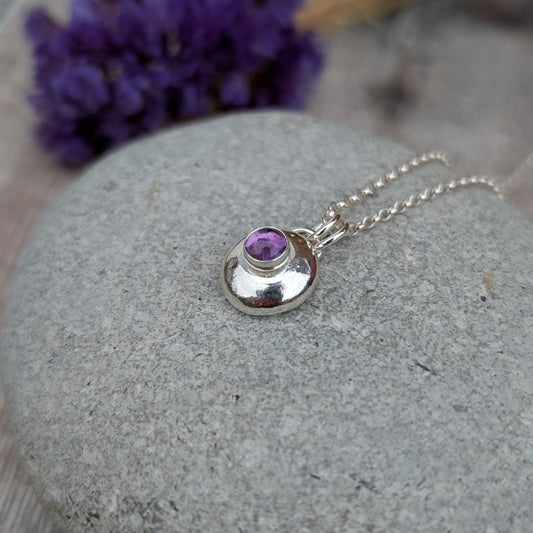 Close-up of a handmade silver pebble necklace with a round amethyst stone, resting on a smooth grey stone with purple flowers blurred in the background.