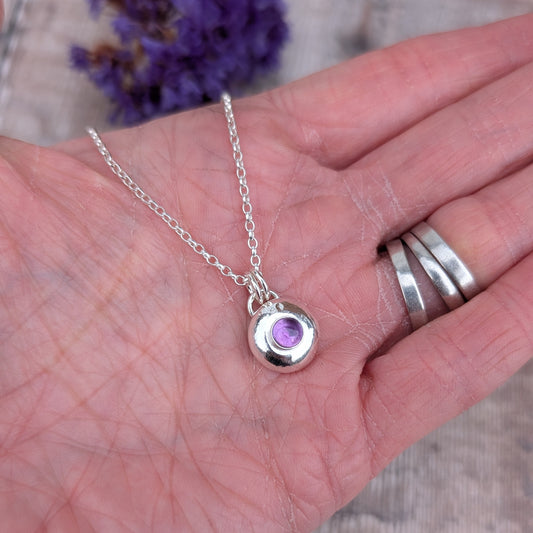 Hand holding a small silver pebble pendant with a purple amethyst set in the centre, showing the scale of the necklace.
