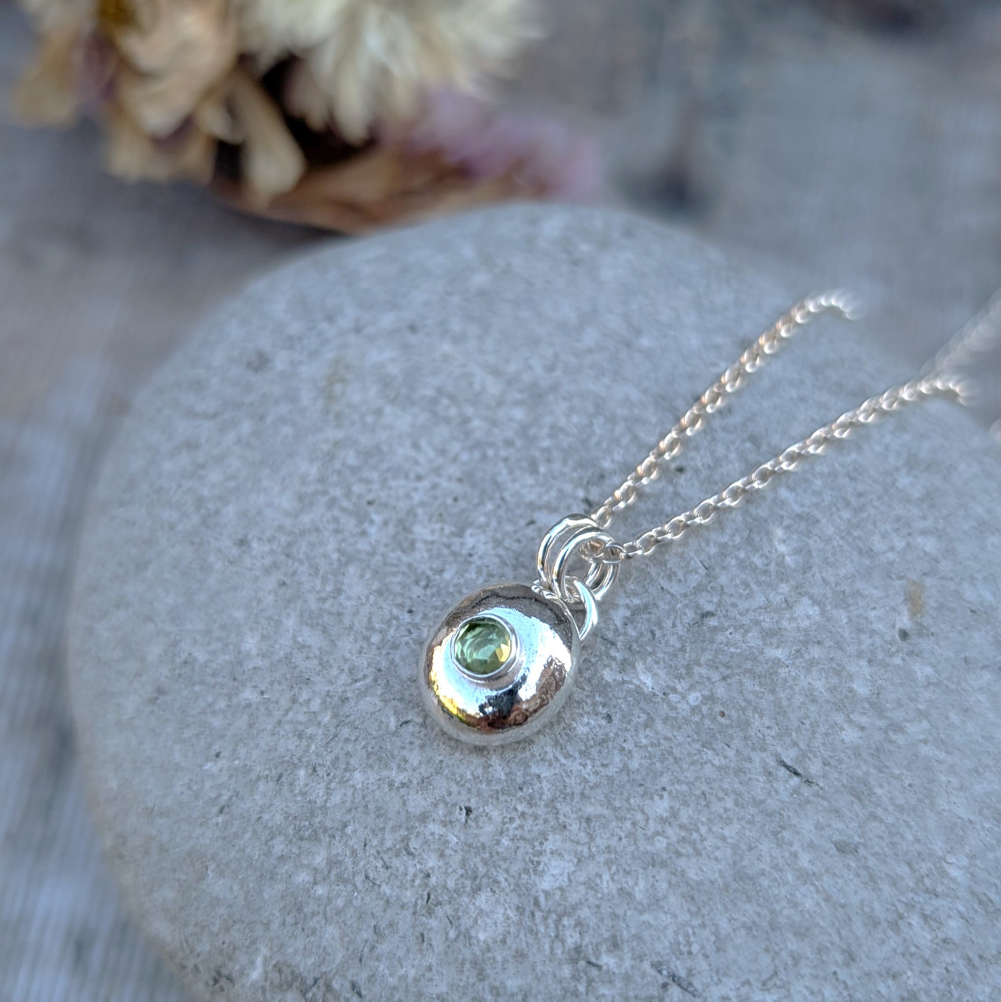 Silver pebble necklace with Peridot pendant close-up on a grey stone, chain draped around.