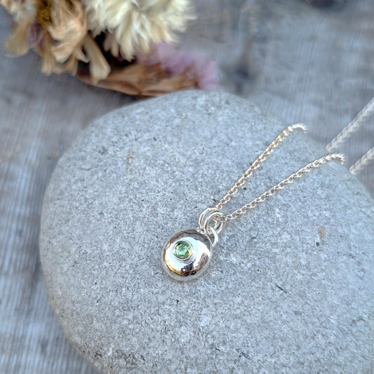 Silver pebble necklace with Peridot pendant resting on a smooth grey stone, dried flowers in the background.