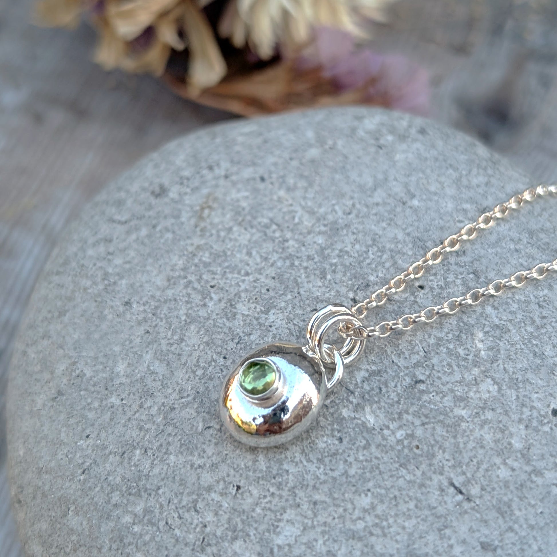 Silver pebble necklace with Peridot pendant shown from above on a grey stone surface.