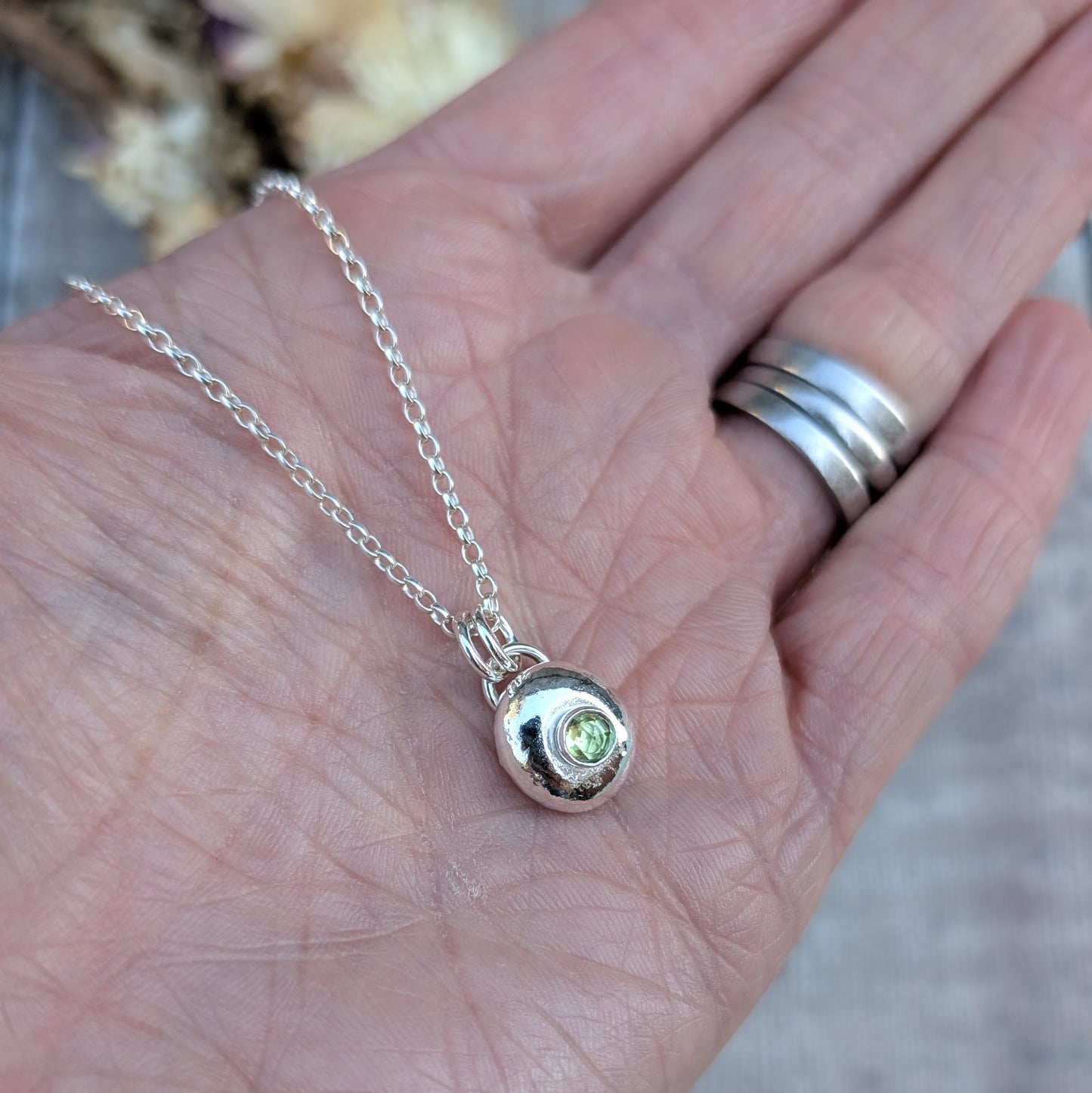 Silver pebble necklace with Peridot pendant resting on a hand with silver rings.