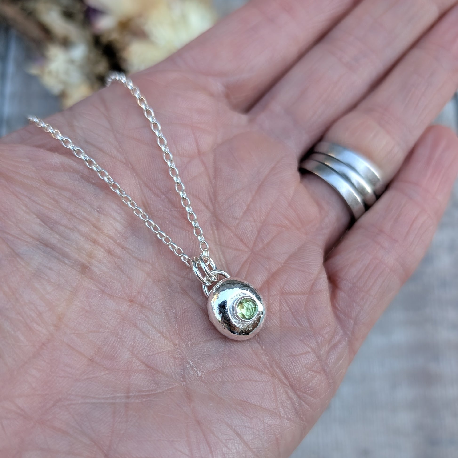 Silver pebble necklace with Peridot pendant resting on a hand with silver rings.