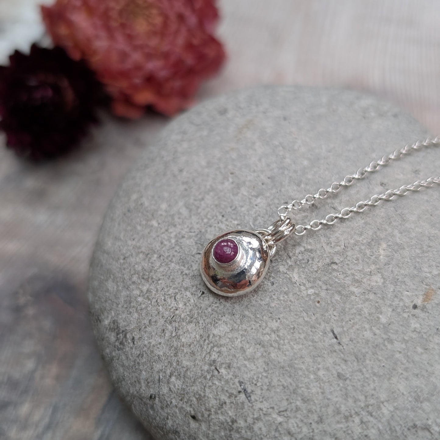 Silver pebble necklace with ruby pendant resting on a stone, surrounded by red dried flowers.