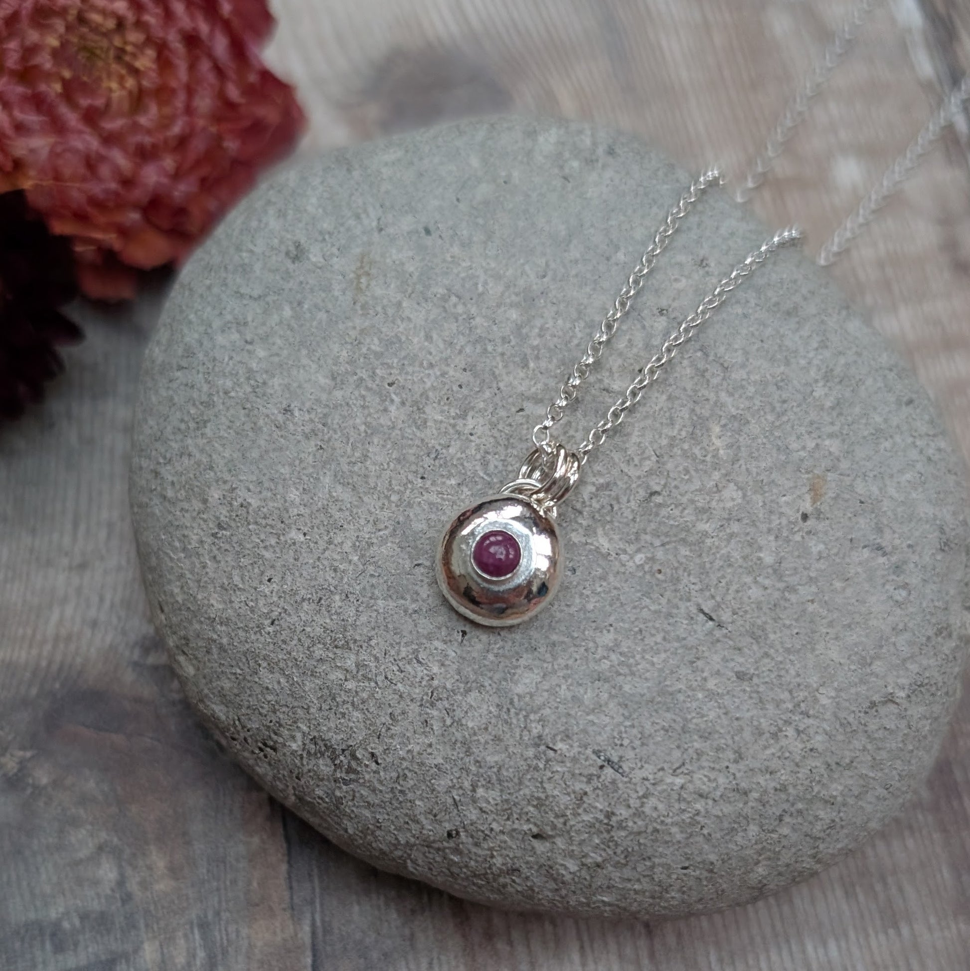 Silver pebble necklace with ruby pendant resting on a smooth grey stone with dried flowers in the background.