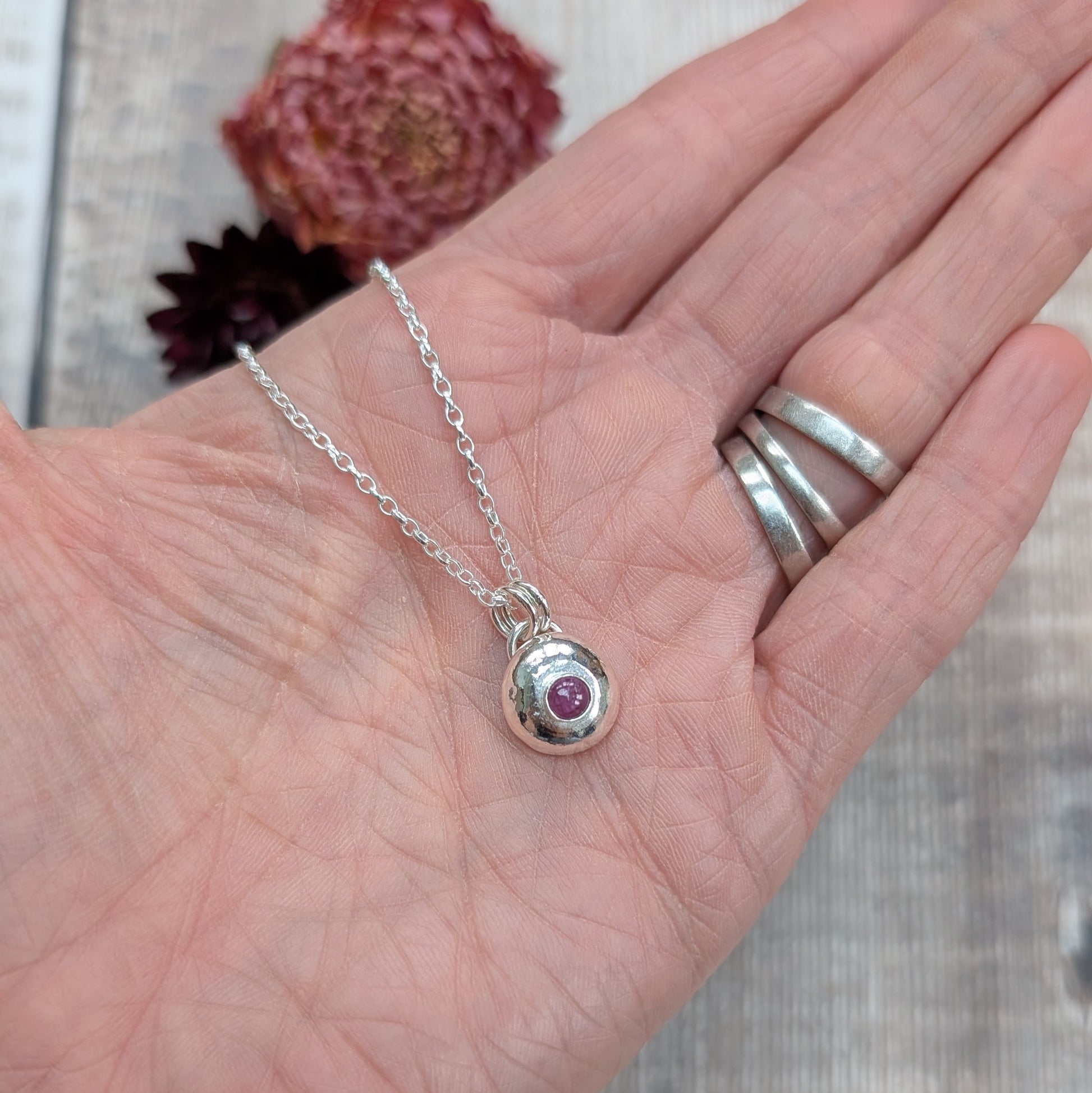 Silver pebble necklace with ruby pendant displayed on an open hand, highlighting the small round pebble design.
