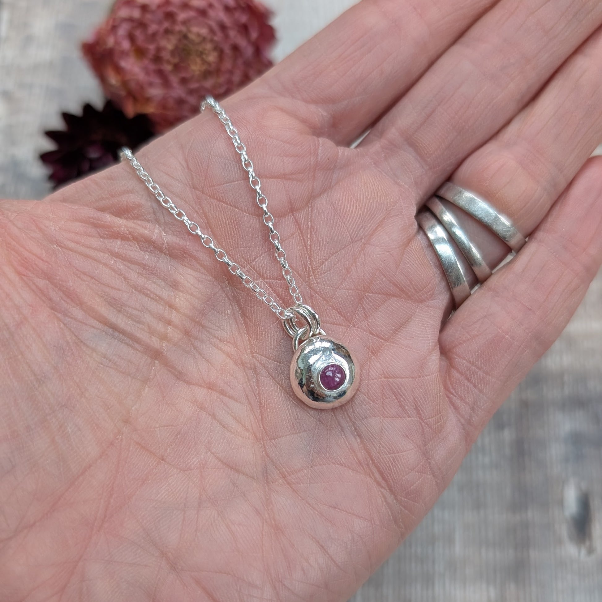 Another view of the silver pebble necklace with ruby pendant shown in a hand, with flowers softly blurred in the background.