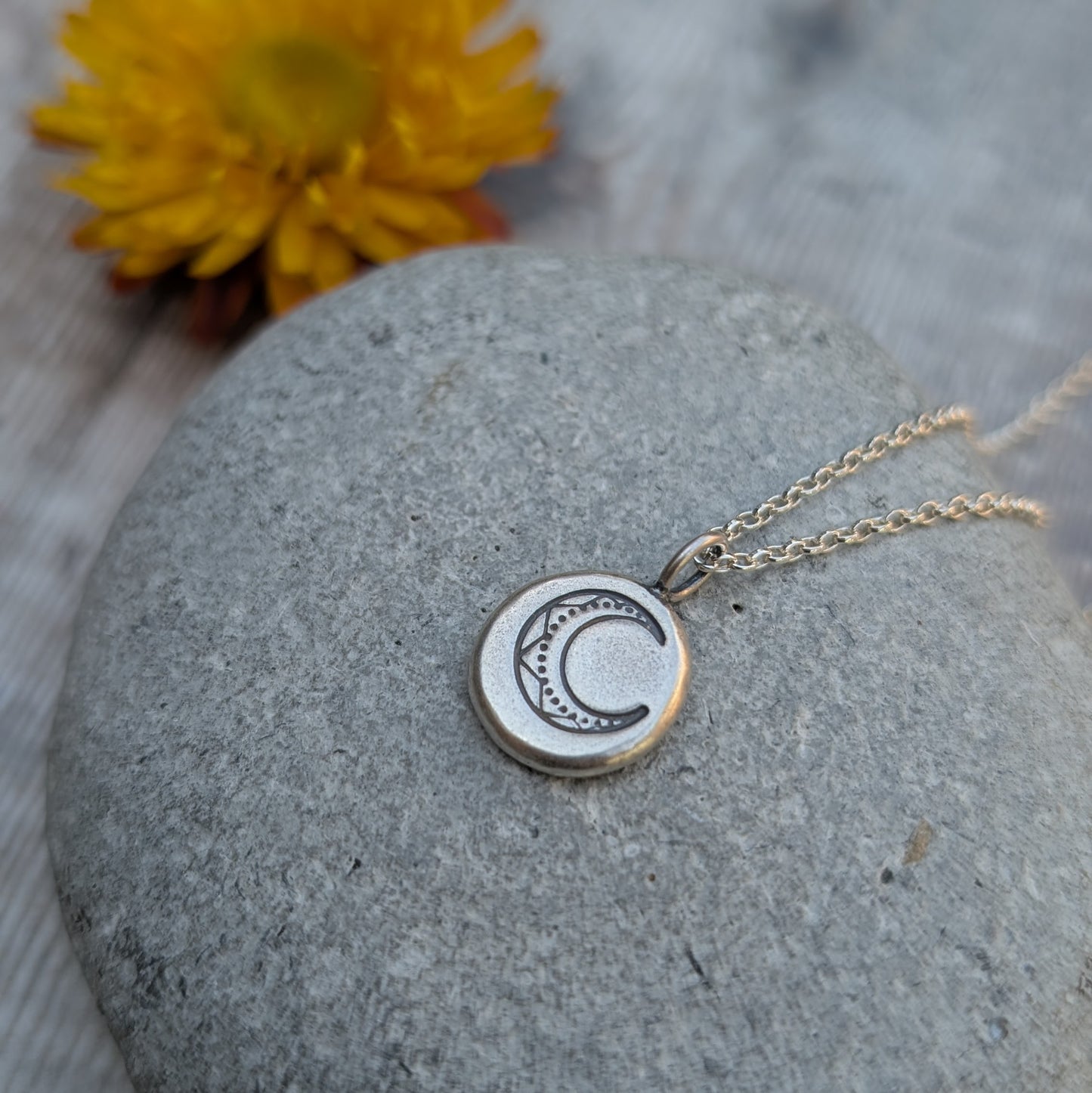 Silver Pebble Stamped Moon Necklace draped across a stone with a yellow flower blurred in the background.