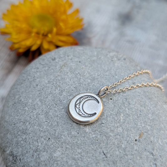 Silver Pebble Stamped Moon Necklace displayed on a smooth stone with a yellow flower in the background.