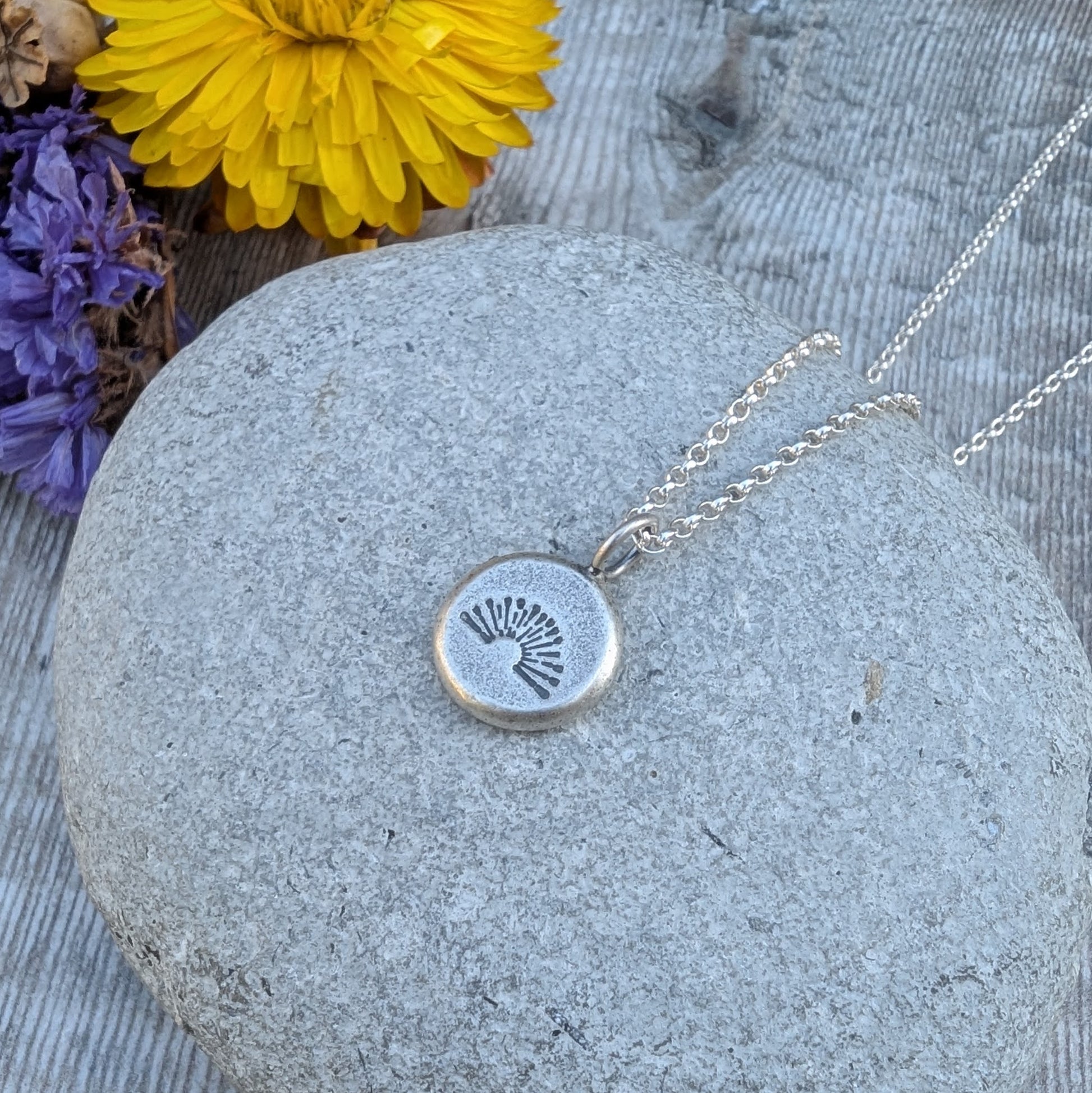 Close-up of silver pebble stamped sunburst necklace on a stone with flowers around it.