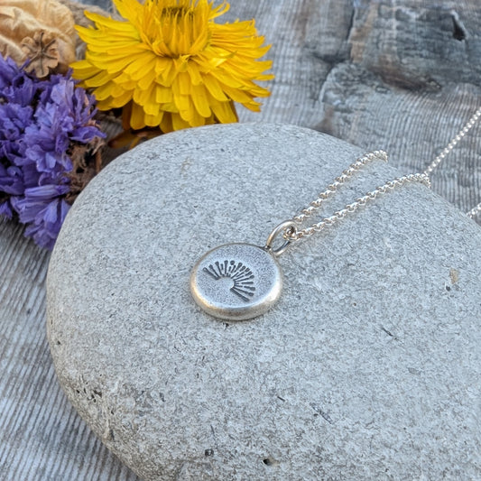 Silver pebble stamped sunburst necklace resting on a stone with yellow and purple flowers in the background.