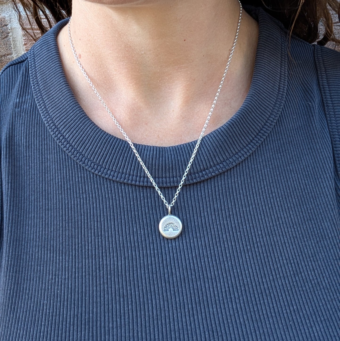 Silver pebble stamped sunburst necklace worn on a chain against a dark top.