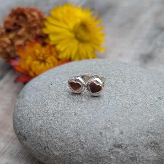 Silver Pebble Stud Earrings with Copper Hearts resting on a smooth grey stone, with yellow and orange flowers softly blurred in the background.