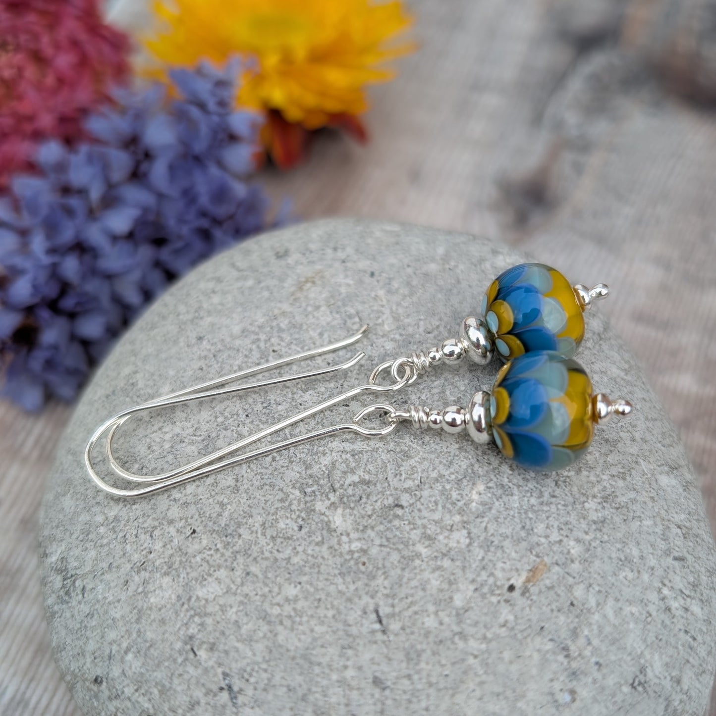 Silver petals lampwork glass bead earrings displayed on a smooth grey stone, featuring round handmade glass beads with blue and mustard yellow petal patterns and long silver ear wires.