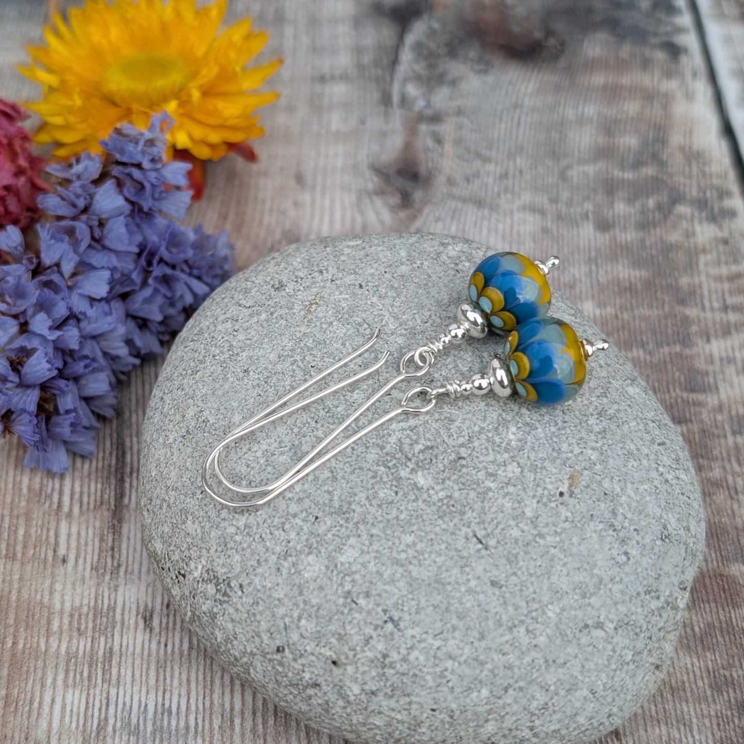 Silver petals lampwork glass bead earrings resting on a grey stone with dried flowers in the background, showing detailed blue and yellow petal designs in the glass beads.
